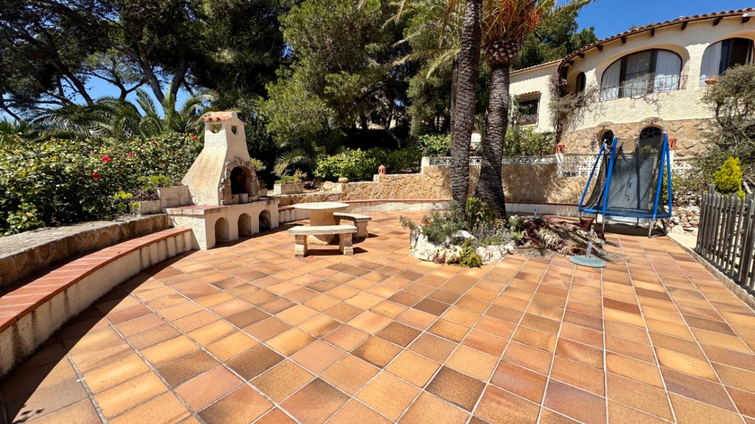 Amplia terraza de baldosas de terracota con barbacoa de piedra integrada, mobiliario de piedra y palmeras junto a una villa de estilo mediterráneo.
