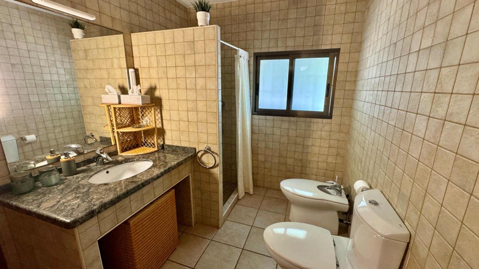 Baño con encimera de granito, lavabo integrado y accesorios de mimbre. Incluye paredes alicatadas en beige, ducha de obra con cortina y zona de sanitario con bidé.