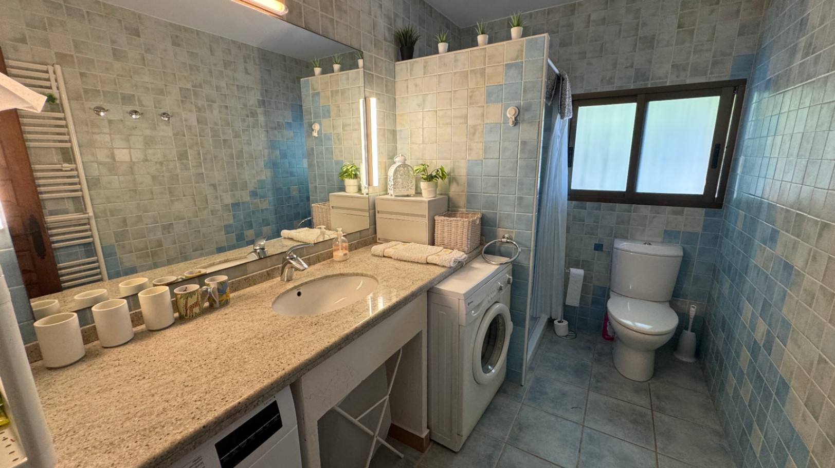 Baño con encimera de granito, paredes de mosaico azul y lavadora integrada. Dispone de un gran espejo con iluminación superior y ventana exterior.