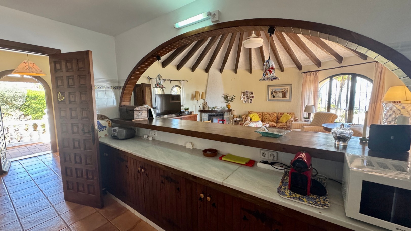 Barra de cocina con encimera de mármol y muebles de madera, abierta mediante un gran arco hacia un salón circular con vigas radiales y chimenea.