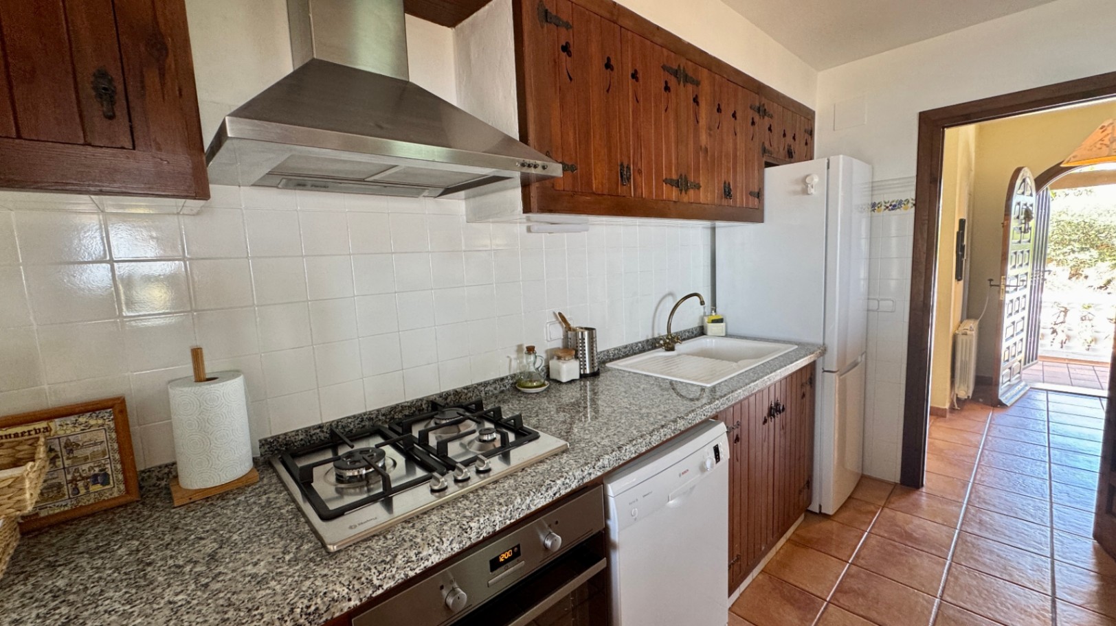 Cocina de estilo rústico con armarios de madera oscura, encimera de granito y placa de gas. Incluye suelos de terracota, salpicadero de azulejos blancos y electrodomésticos.