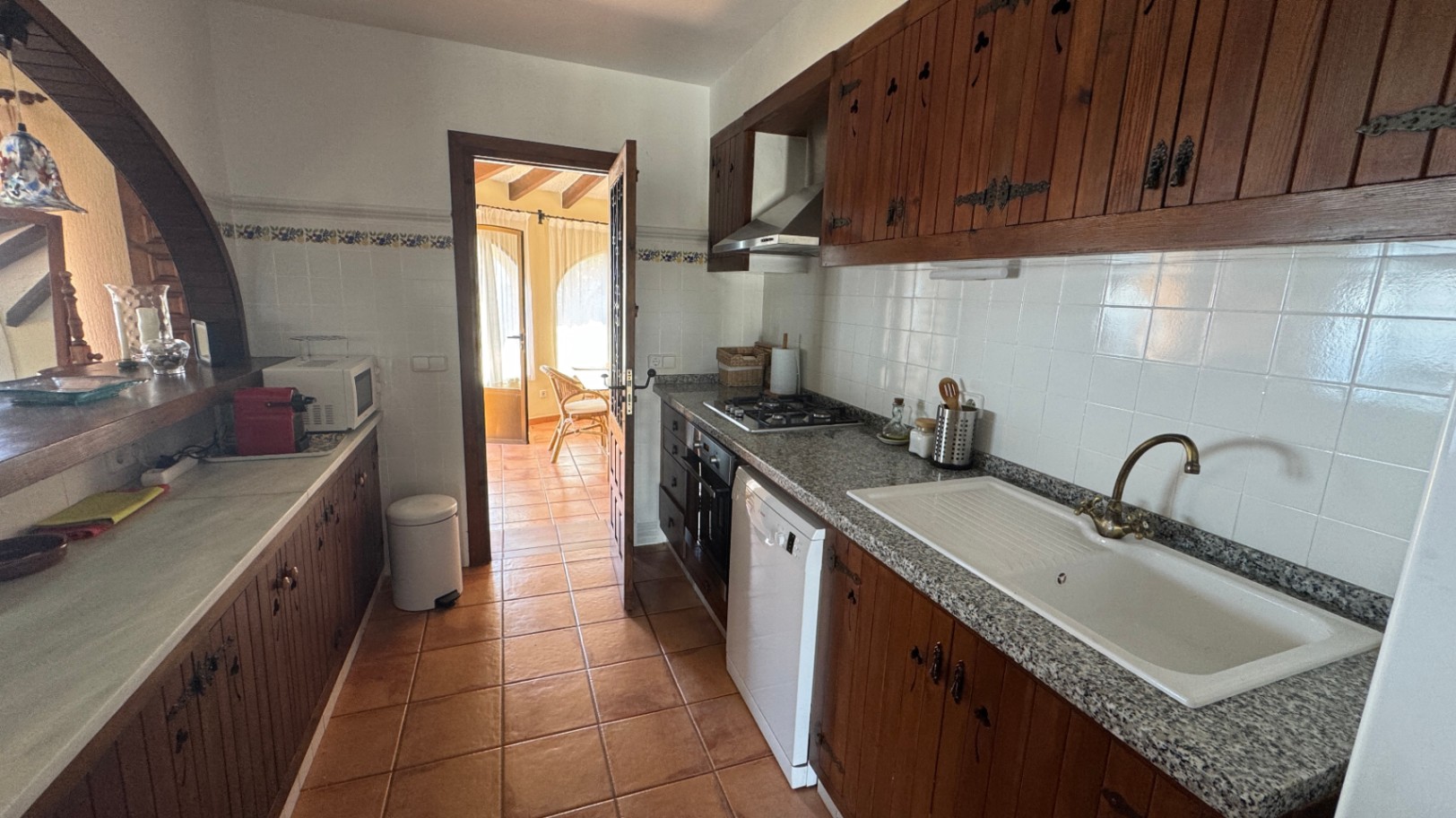 Cocina rústica con armarios de madera oscura, encimeras de granito y suelos de terracota. Dispone de zona de cocción a gas y un arco pasaplatos lateral.