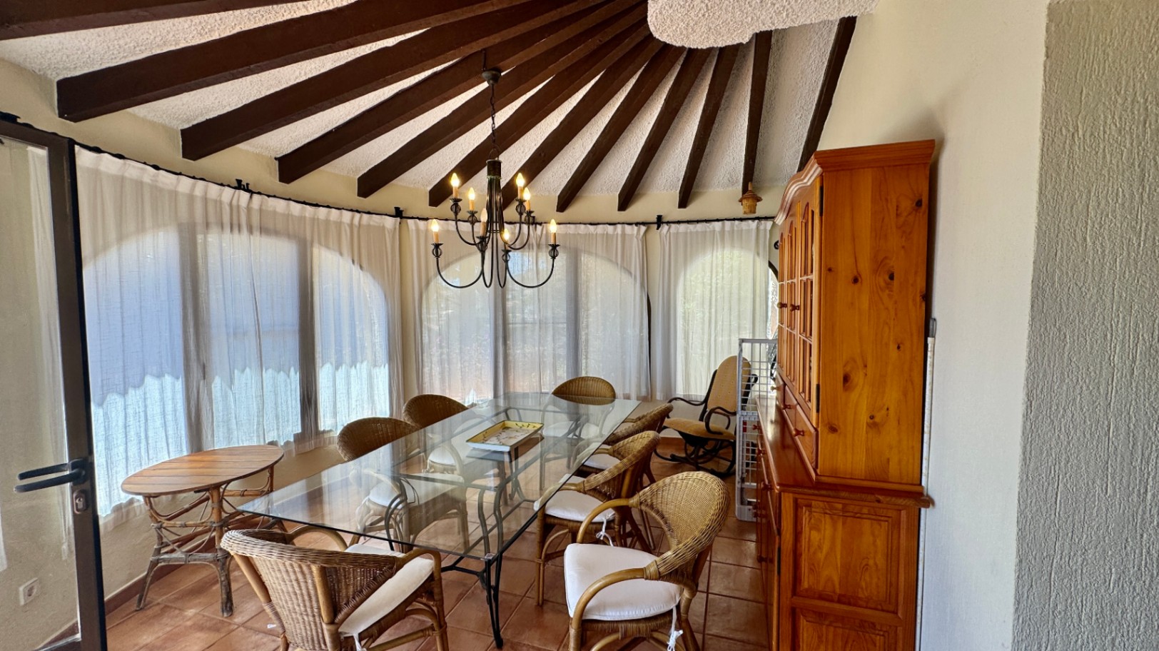 Comedor con techo de vigas de madera radiales, mesa de cristal con sillas de mimbre y una alacena de madera clásica sobre suelo de terracota.