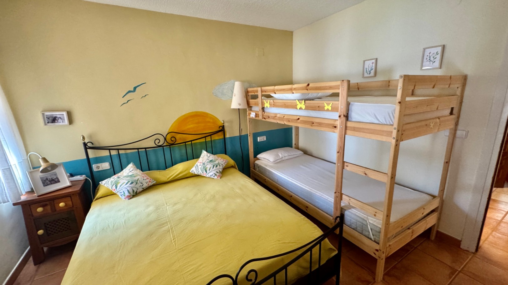 Dormitorio con cama doble de forja negra y litera de madera clara. La estancia dispone de suelos de terracota y una pared con un mural decorativo de sol y mar.