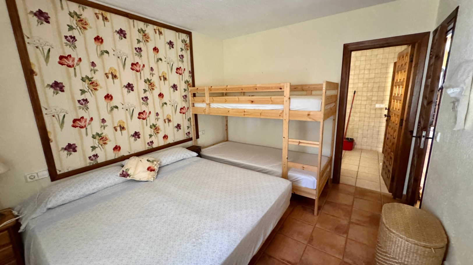 Dormitorio con cama doble y literas de madera, destacado por un panel de papel pintado floral y suelos de terracota. Incluye acceso directo a un baño alicatado.