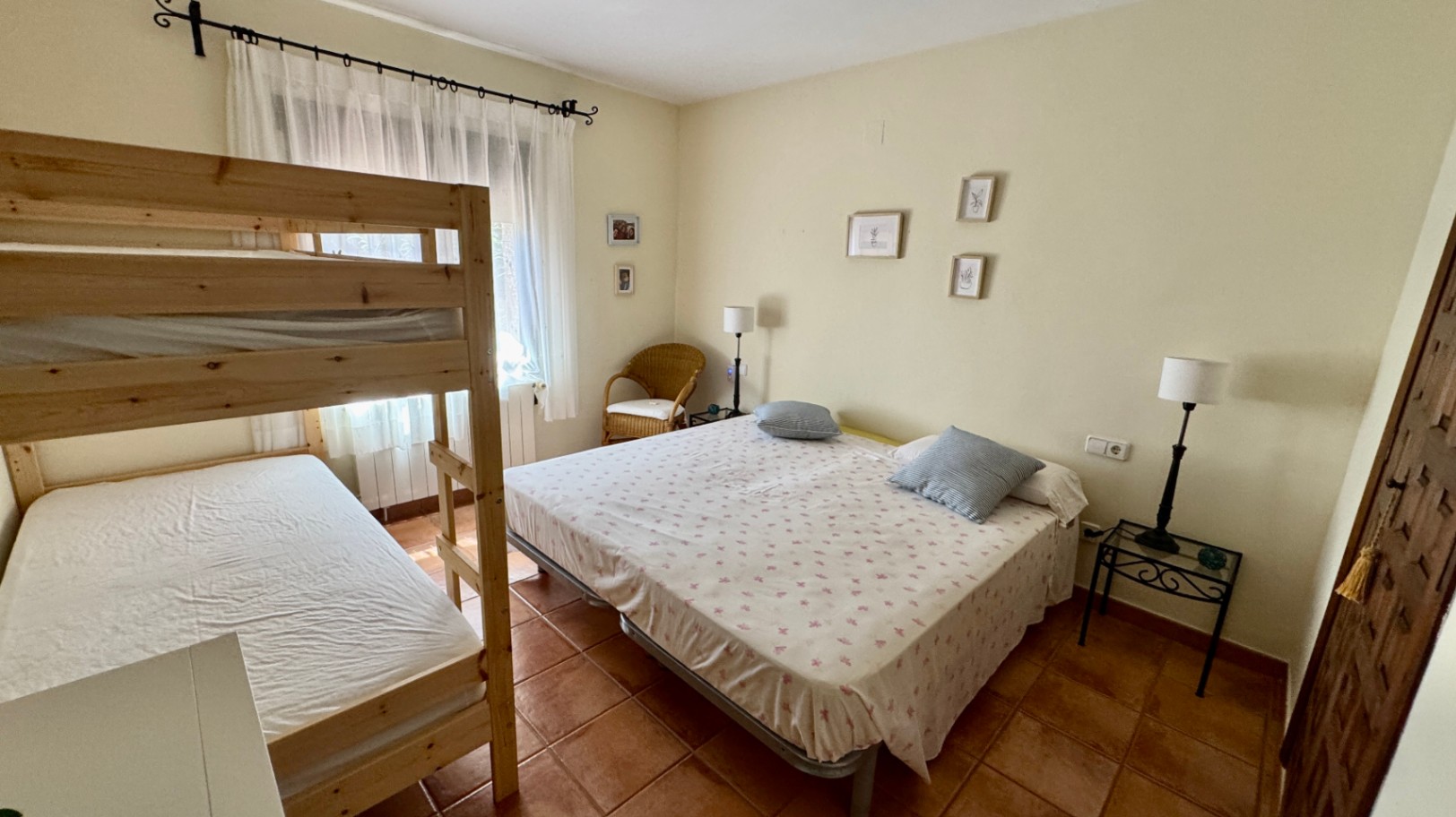 Dormitorio con literas de madera y cama doble, suelo de baldosas de barro cocido y luz natural. Incluye un sillón de mimbre y decoración sencilla en tonos neutros.