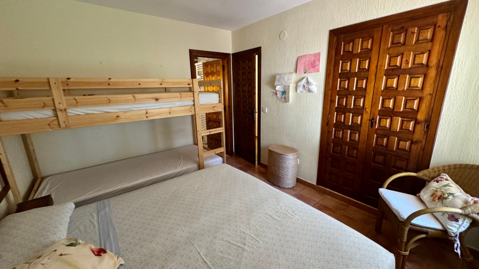 Dormitorio con literas de pino, armario empotrado de madera con cuarterones y suelos de terracota. Incluye un sillón de mimbre y paredes con acabado rugoso.