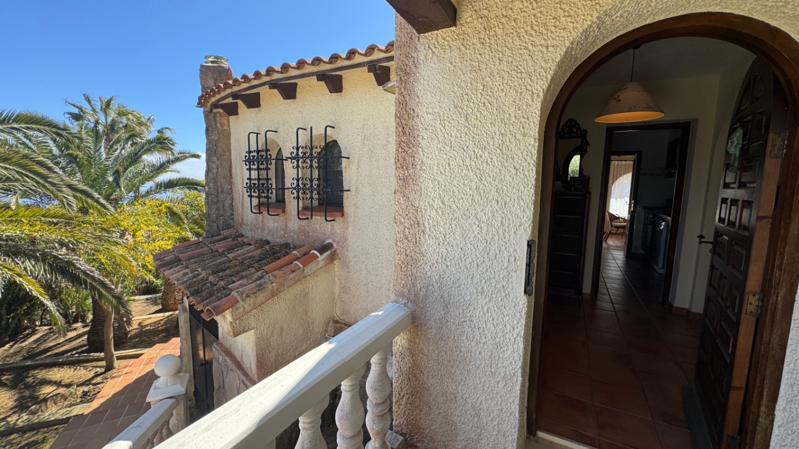 Entrada principal arqueada con puerta de madera maciza, suelos de terracota y ventanas con rejas de hierro forjado bajo un cielo despejado.
