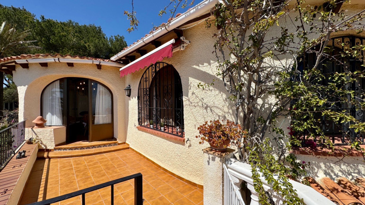 Exterior de villa mediterránea con terraza de baldosas de terracota, ventanas en arco con rejas de hierro forjado y un toldo retráctil rojo.