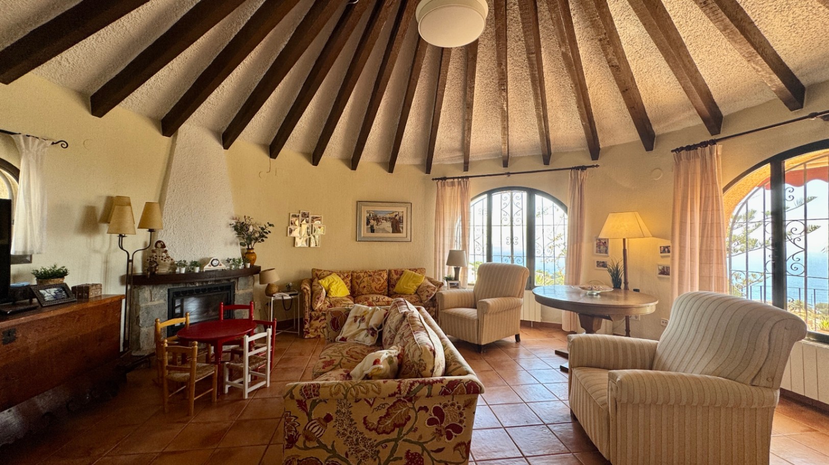 Salón circular con techo cónico de vigas de madera radiales, suelo de terracota, chimenea de piedra y ventanales en arco con vistas al mar.