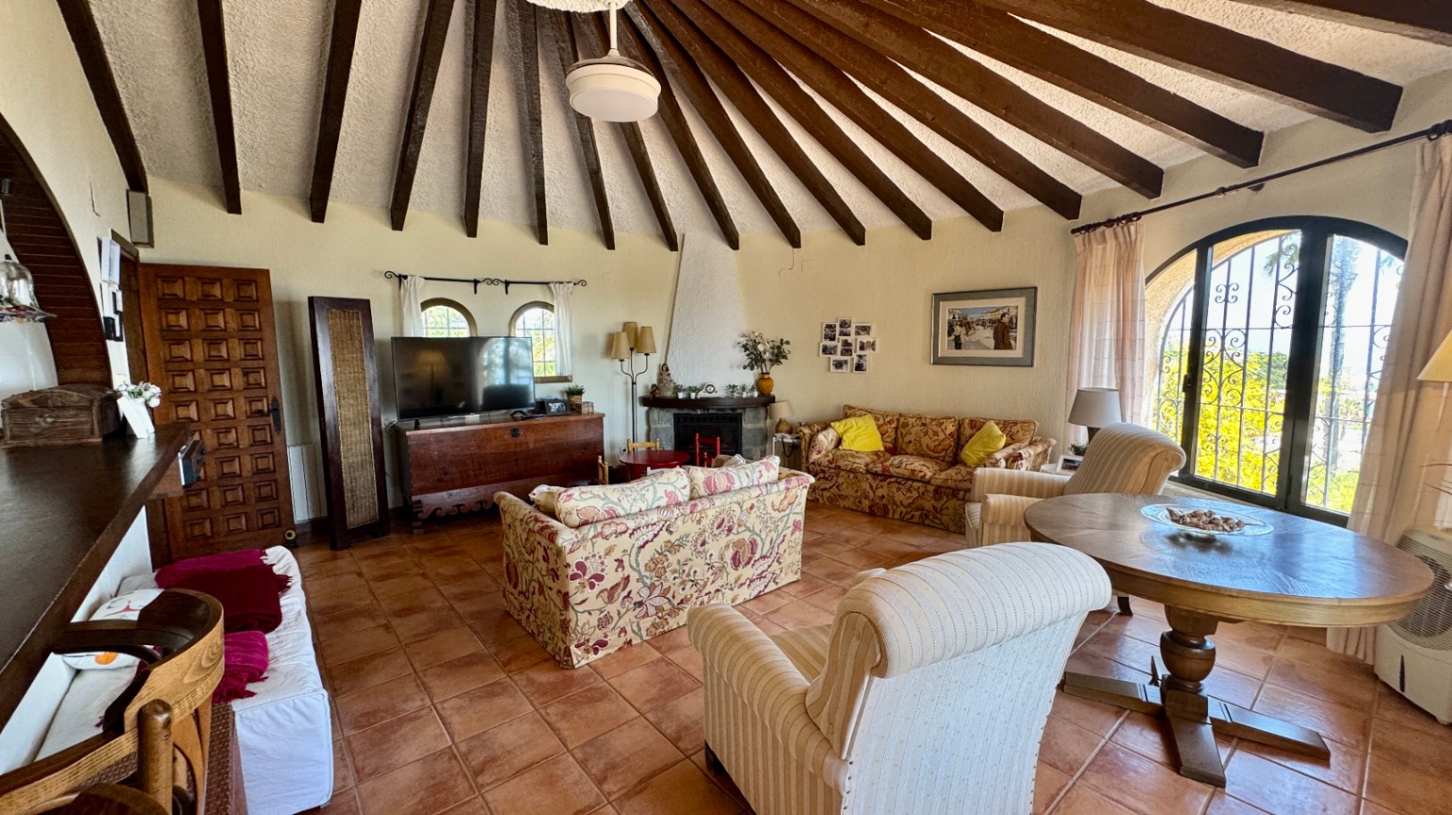 Salón con techo abovedado de vigas de madera radiales, suelos de terracota, chimenea y ventanales en arco con rejería artesanal.