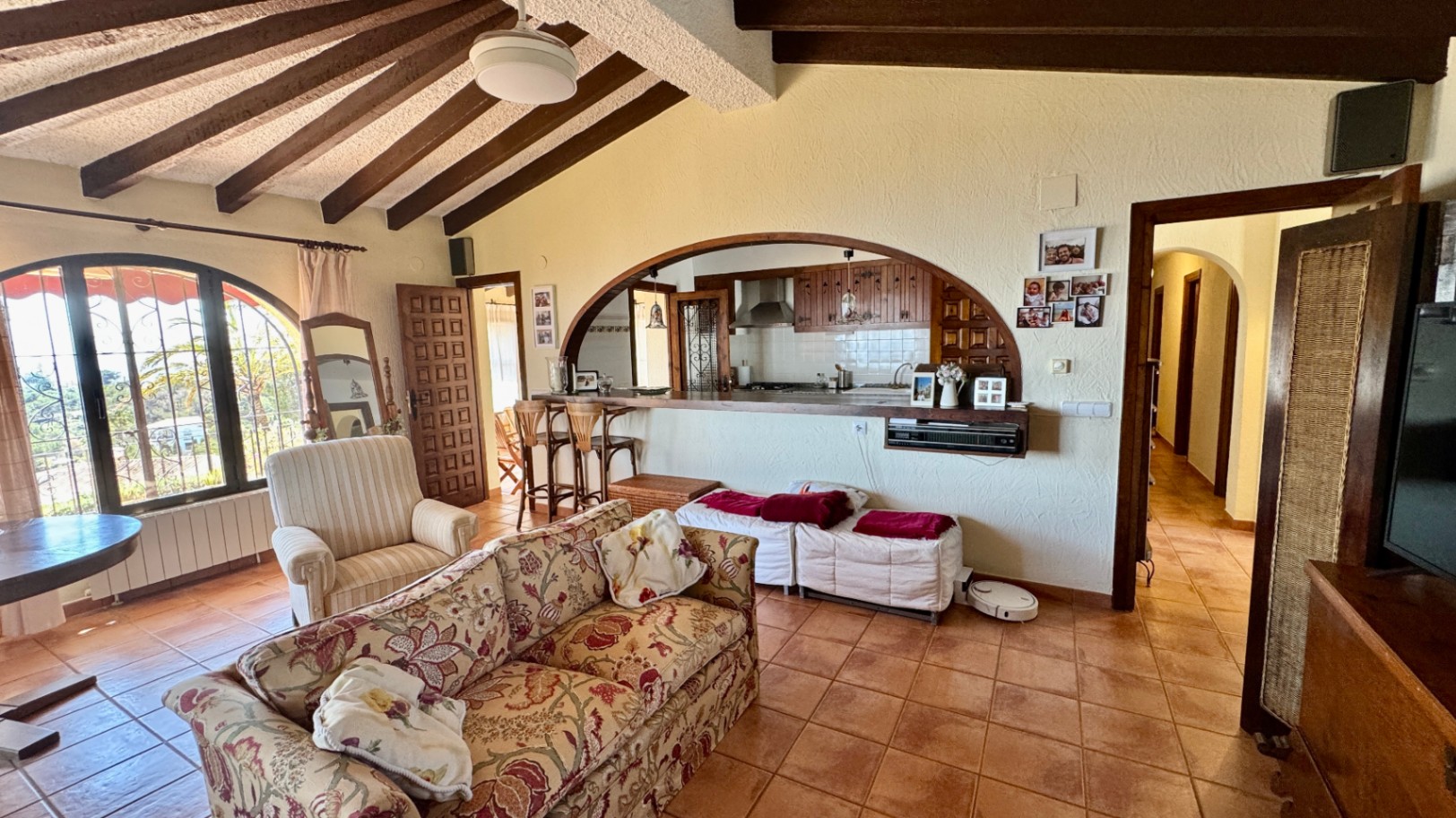 Salón con techos inclinados de vigas de madera oscura, suelos de terracota y un gran arco que comunica con la cocina a través de una barra de madera.