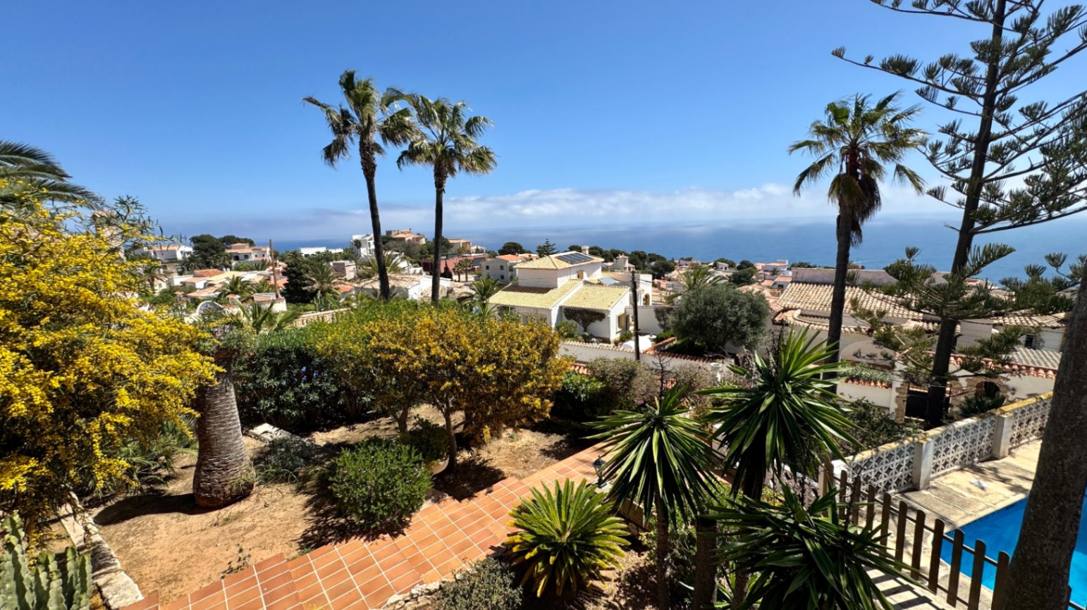 Vista elevada de un jardín mediterráneo con senderos de terracota y palmeras, con vistas a una zona residencial costera y al mar Mediterráneo.