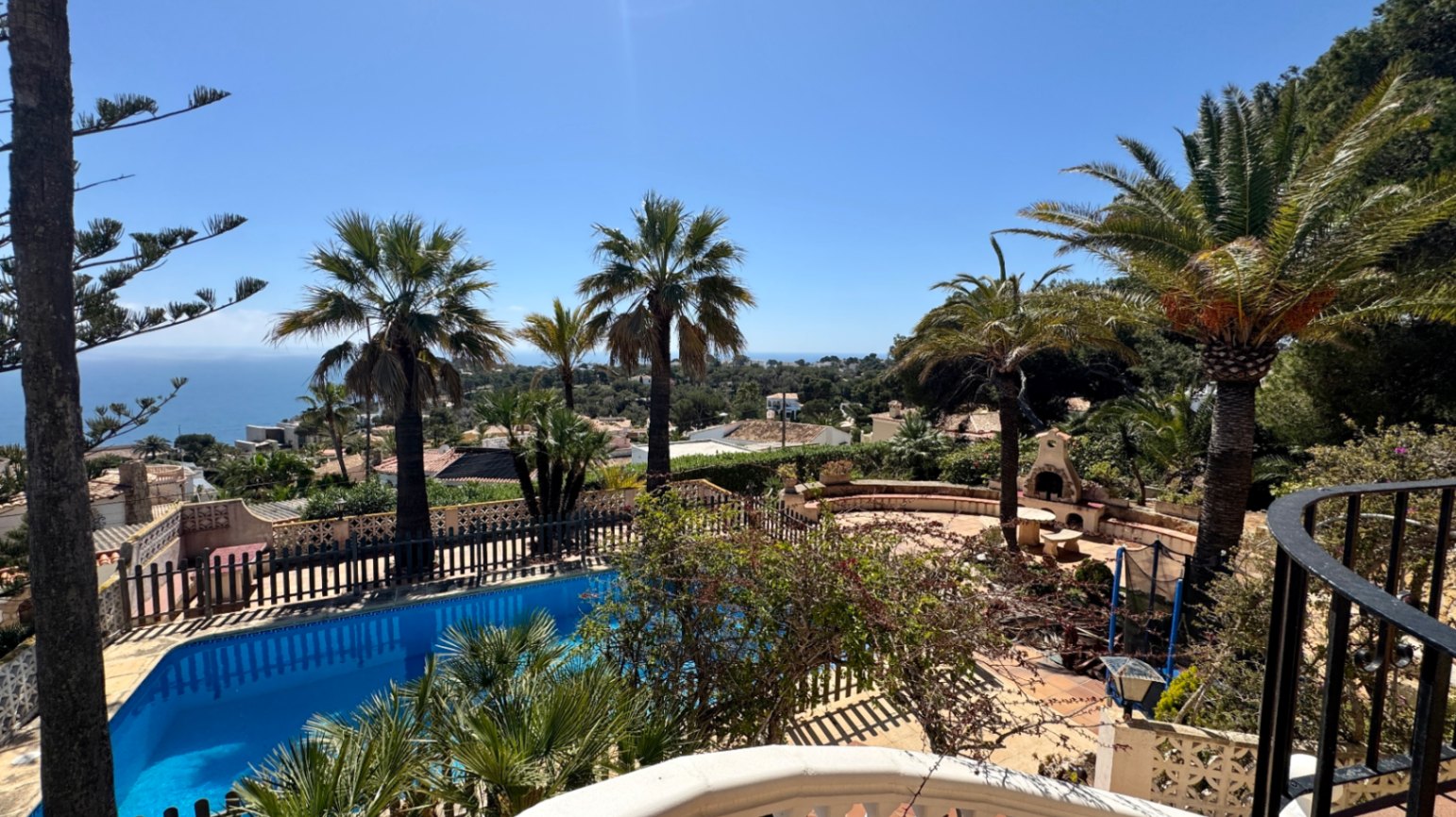 Vista elevada de una piscina privada y patio de piedra con chimenea exterior, rodeado de palmeras y vistas panorámicas al mar Mediterráneo.