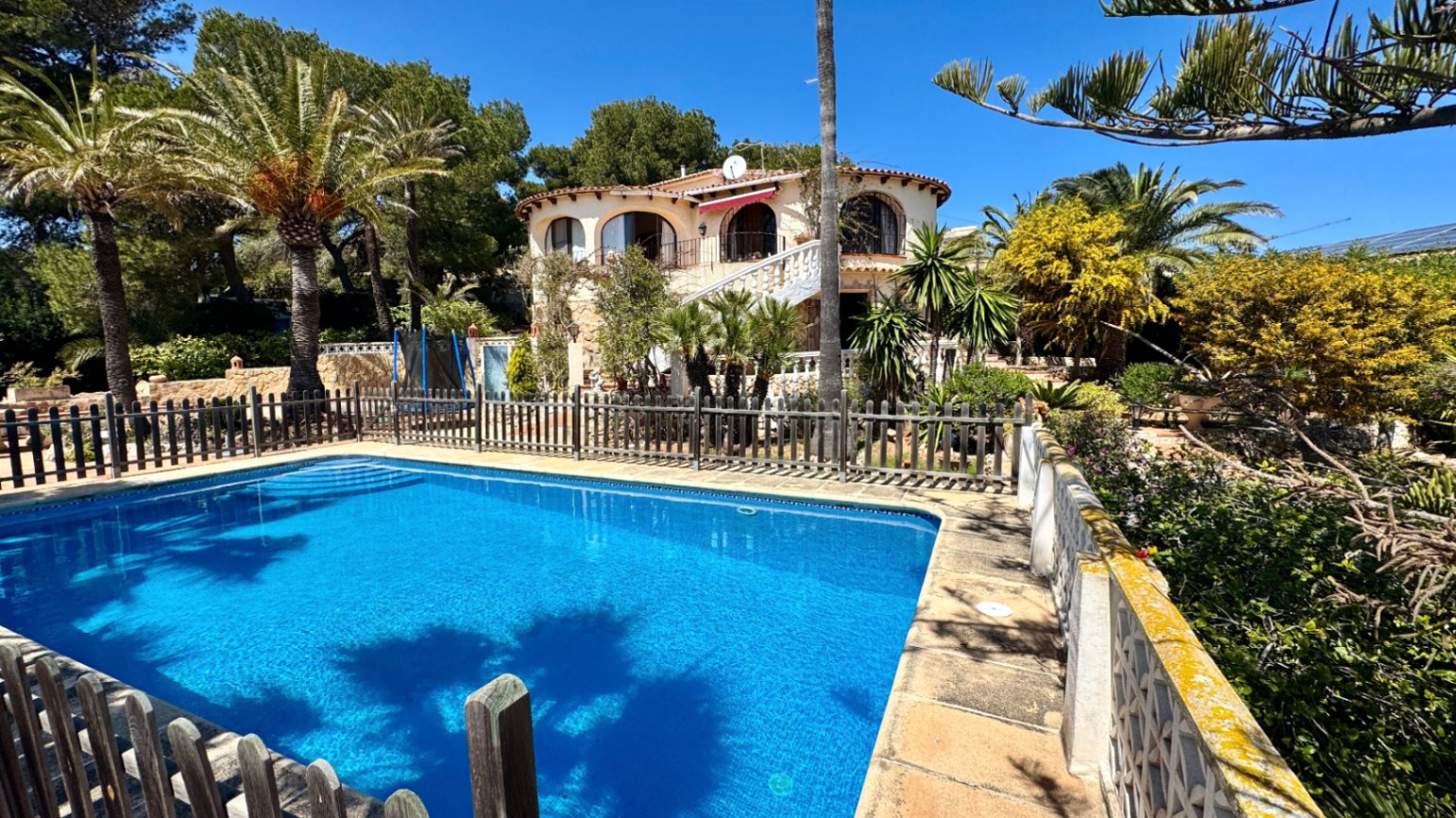 Vista exterior con piscina rectangular vallada y villa de estilo mediterráneo con arcos, rodeada de palmeras y vegetación autóctona.
