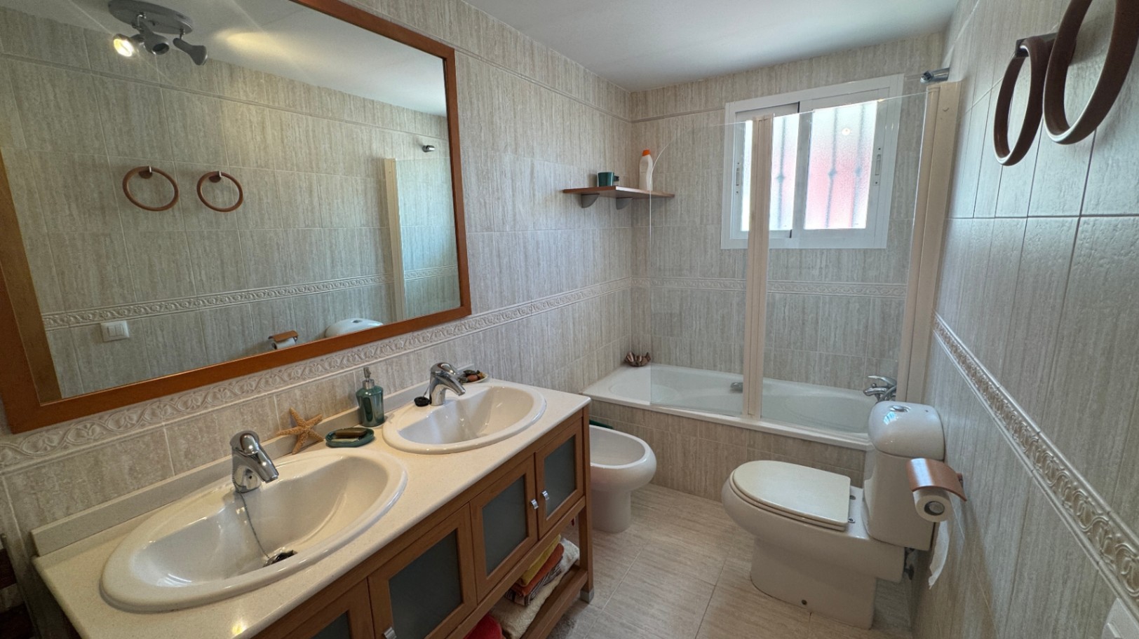 Baño completo con lavabo doble sobre mueble de madera, bañera con mampara de cristal y paredes revestidas en azulejos de tonos neutros con cenefa.