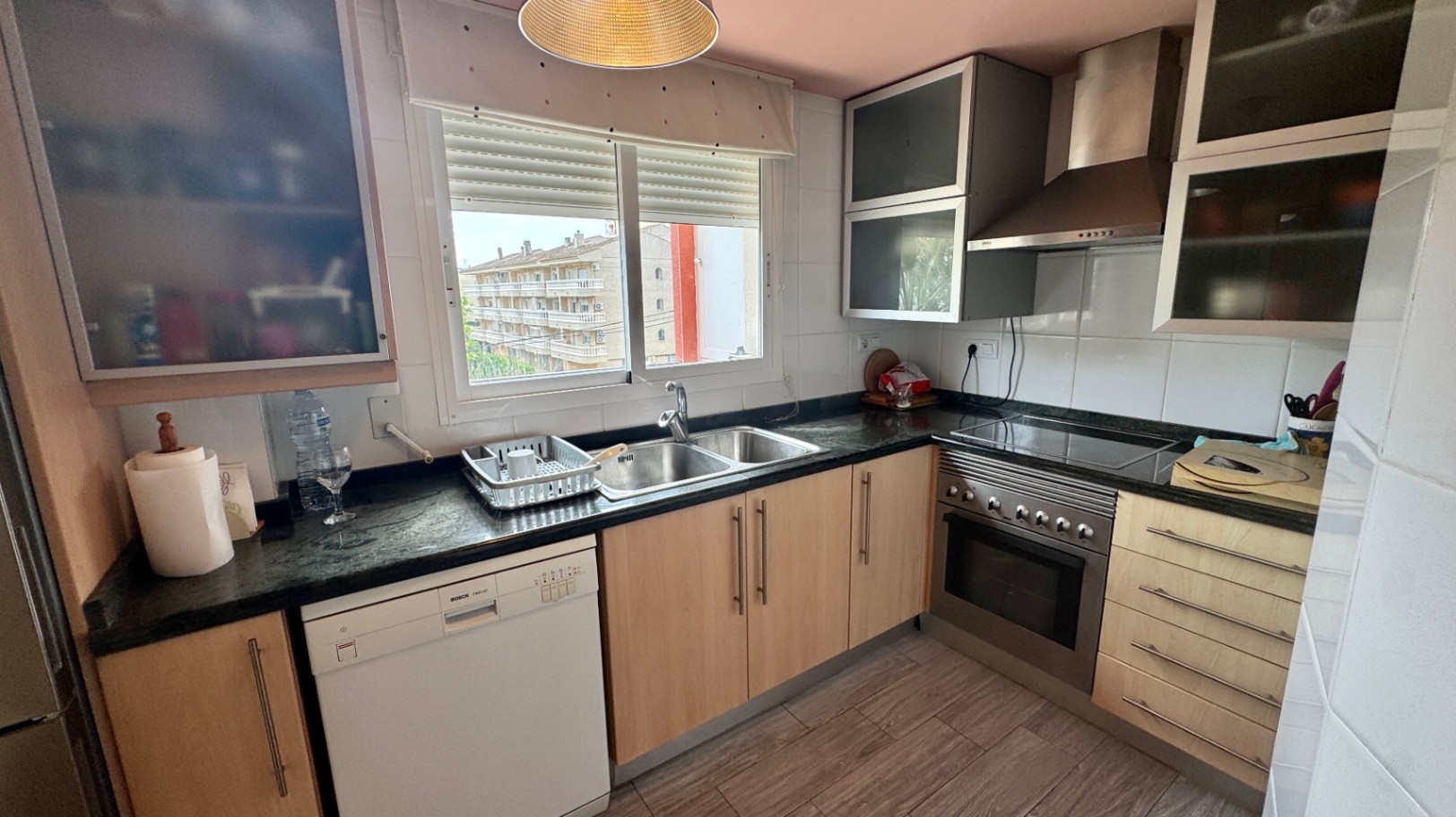 Cocina equipada con muebles de madera clara, encimera de granito oscuro, lavavajillas Bosch y fregadero doble bajo una ventana con vistas exteriores.