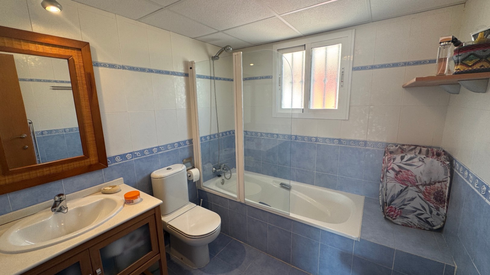 Cuarto de baño con azulejos azules y blancos, bañera con mampara de cristal, espejo con marco de madera y mueble de lavabo a juego.