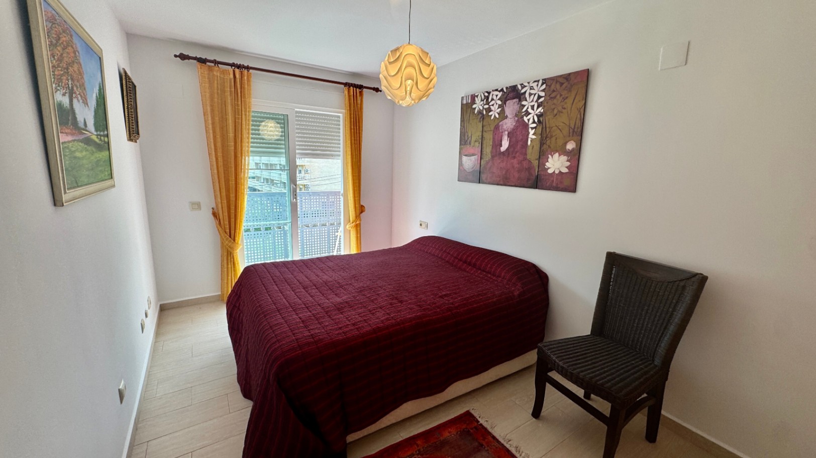 Dormitorio con cama doble, colcha granate y silla de mimbre oscura. Dispone de acceso a un balcón mediante puerta corredera con cortinas amarillas y luz natural.