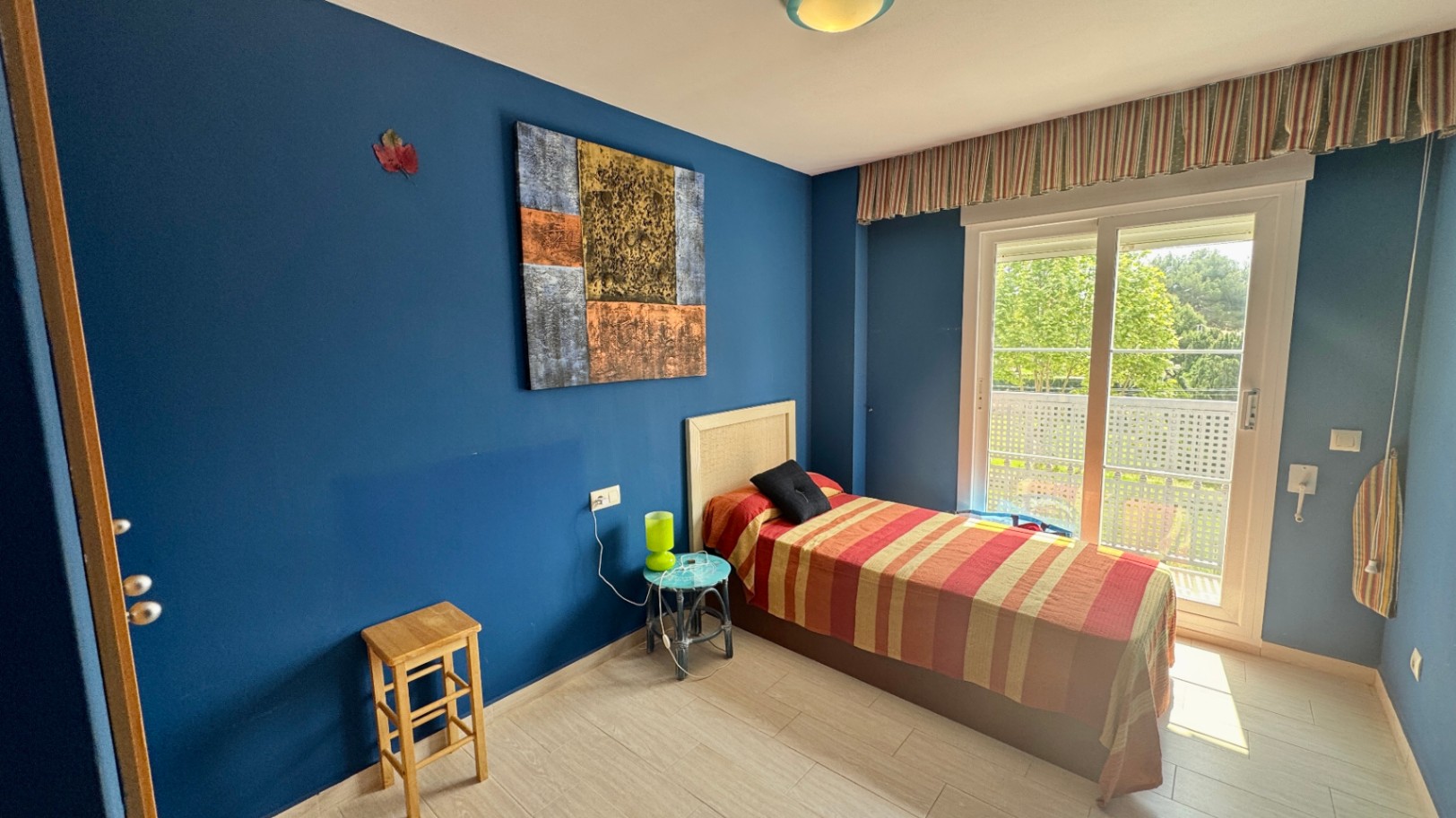 Dormitorio individual con paredes azul intenso, cabecero de mimbre y acceso directo a un balcón privado mediante una gran puerta corredera acristalada.