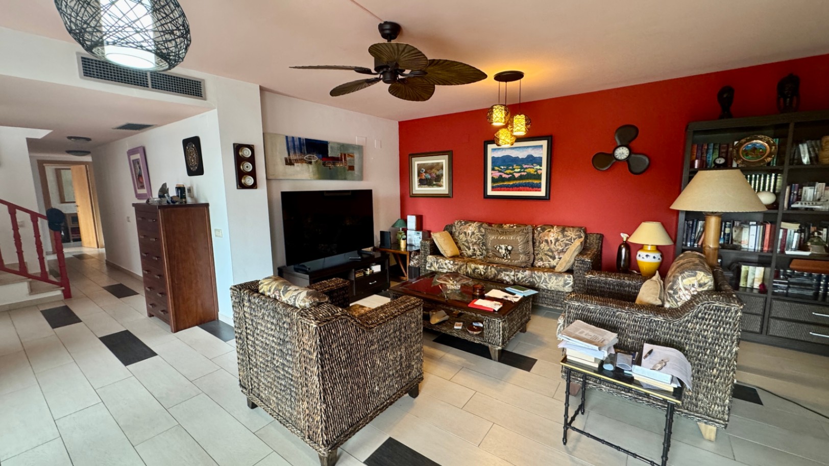 Salón con mobiliario de mimbre, una pared de acento roja con cuadros y ventilador de techo tropical. El suelo cerámico claro incluye detalles oscuros y conecta con el pasillo.
