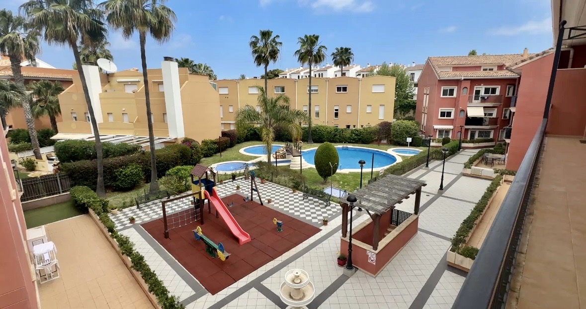 Vista elevada de las zonas comunes con piscina de varios niveles, parque infantil con suelo de caucho y jardines cuidados con palmeras y caminos pavimentados.