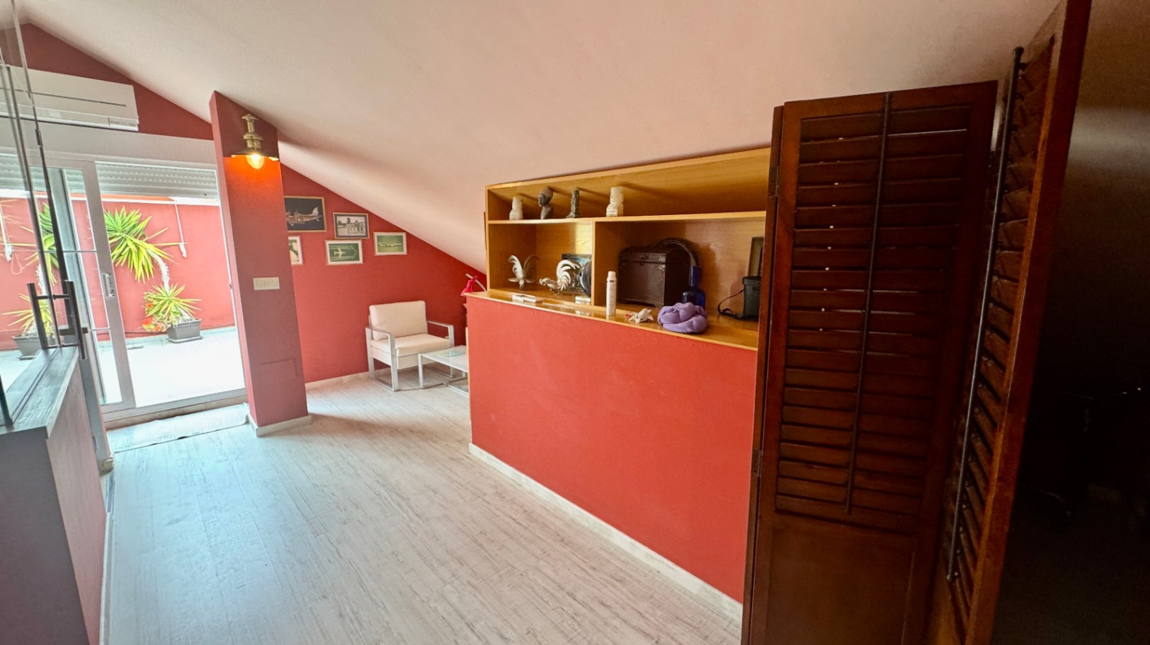 Zona abuhardillada con paredes terracota, techos inclinados y estantería de madera integrada. Dispone de acceso a una terraza privada con plantas mediterráneas.