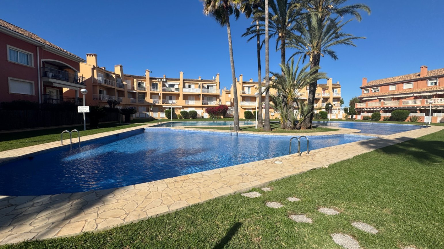 Amplia piscina comunitaria con revestimiento azul, rodeada de un solárium de piedra, césped cuidado y palmeras en un complejo de estilo mediterráneo.