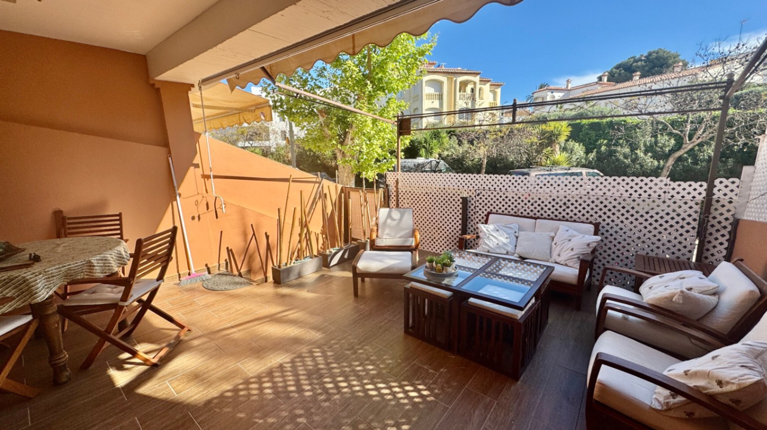 Amplia terraza con suelo cerámico imitación madera, zona de estar con sofás blancos, comedor exterior de madera y toldos extensibles.