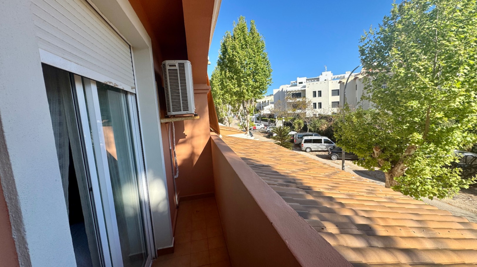 Balcón estrecho con suelo de baldosas de terracota, unidad de aire acondicionado exterior y vistas a una calle arbolada con edificios residenciales.