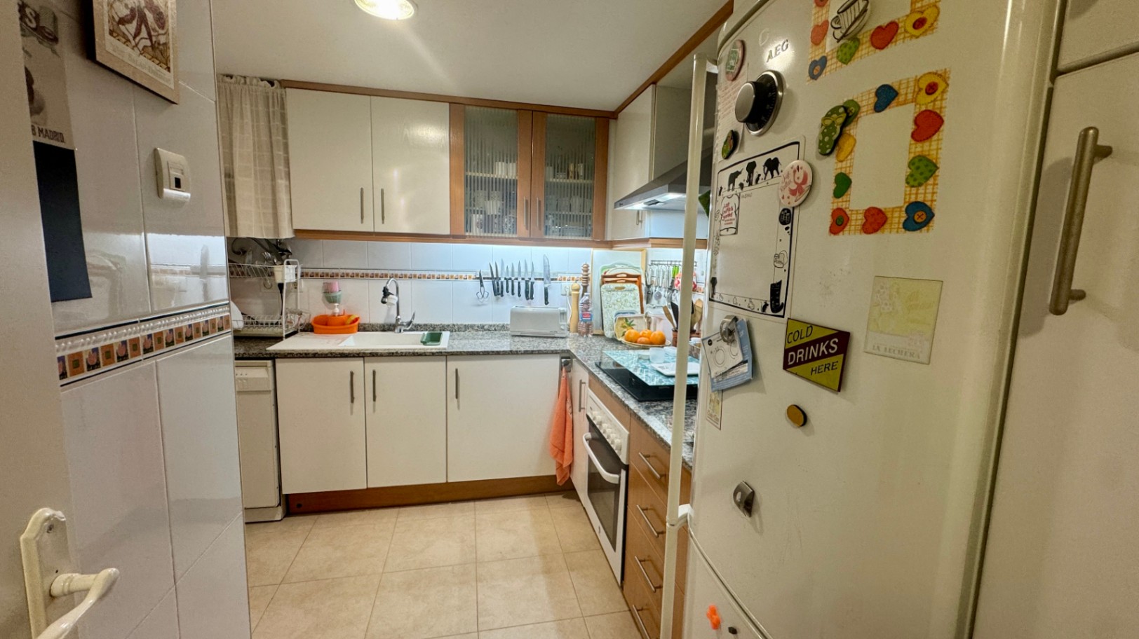 Cocina funcional en forma de L con armarios blancos, encimeras de granito moteado y electrodomésticos integrados bajo una iluminación cálida y dirigida.