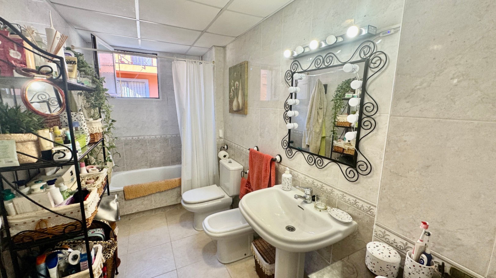 Cuarto de baño alicatado con lavabo de pedestal, espejo de forja con iluminación tipo camerino, bidé y bañera con cortina blanca.