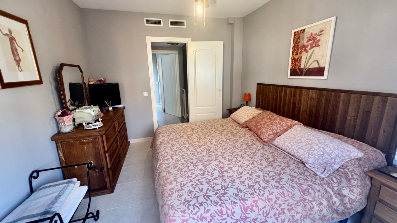 Dormitorio con cabecero de listones de madera oscura, cómoda rústica con espejo y paredes en gris claro. Incluye suelos cerámicos y rejillas de climatización integradas.