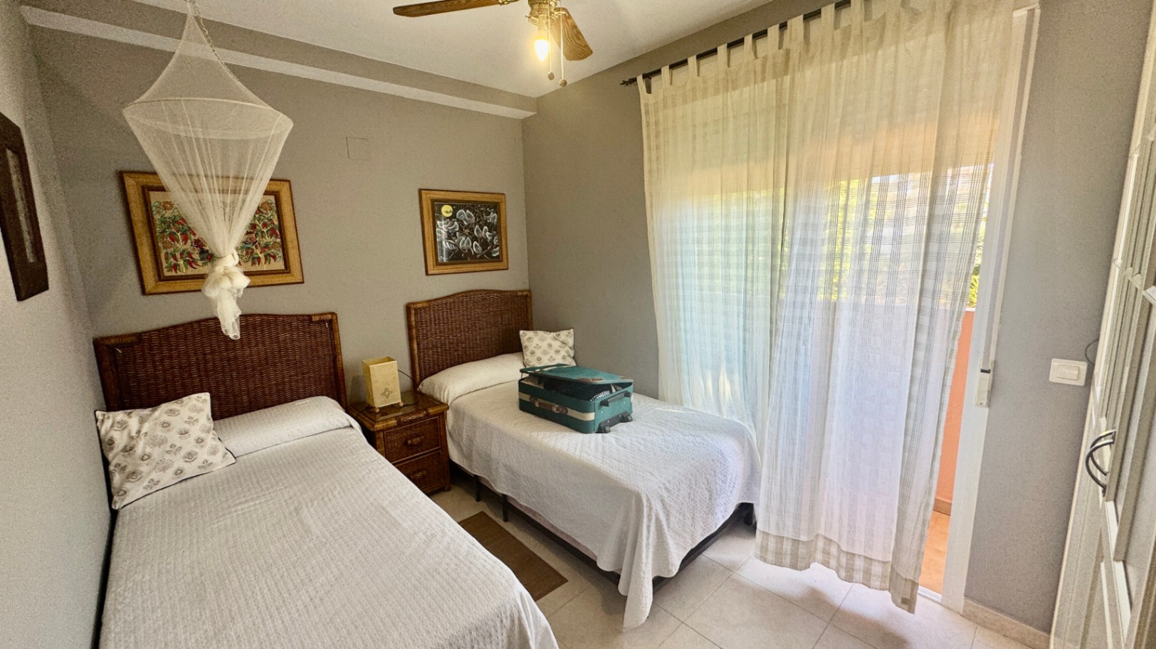 Dormitorio con dos camas individuales, cabeceros de mimbre y acceso a un balcón exterior mediante una puerta acristalada con cortinas ligeras.