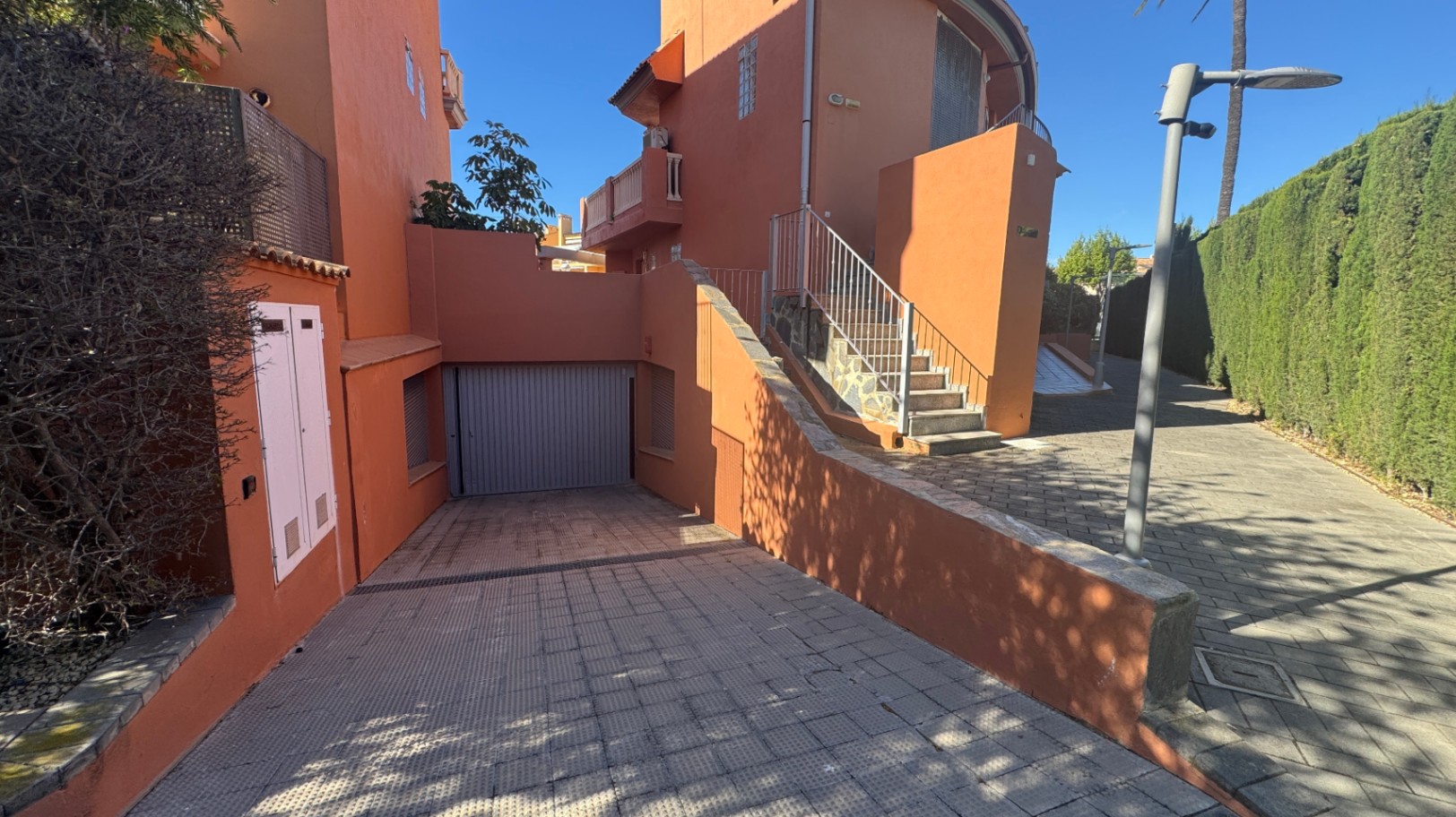 Entrada exterior con rampa pavimentada hacia garaje privado, fachada color terracota y escalera de acceso con barandilla metálica junto a setos altos.