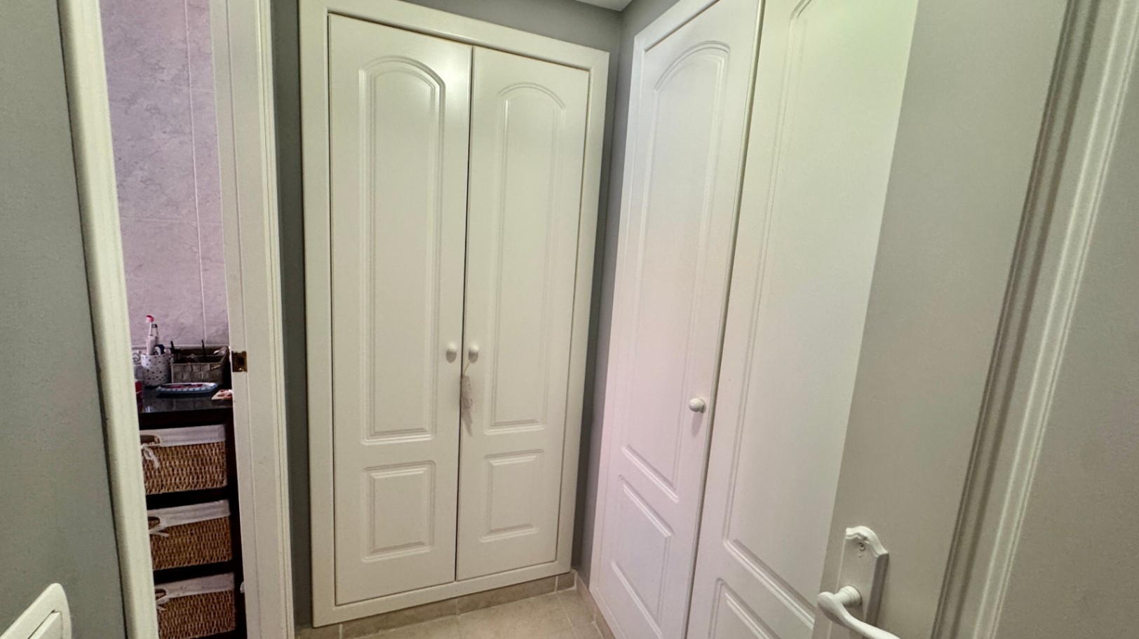 Pasillo con armarios empotrados blancos de suelo a techo, con paneles arqueados y pomos redondos, junto a un baño con cestas de mimbre.