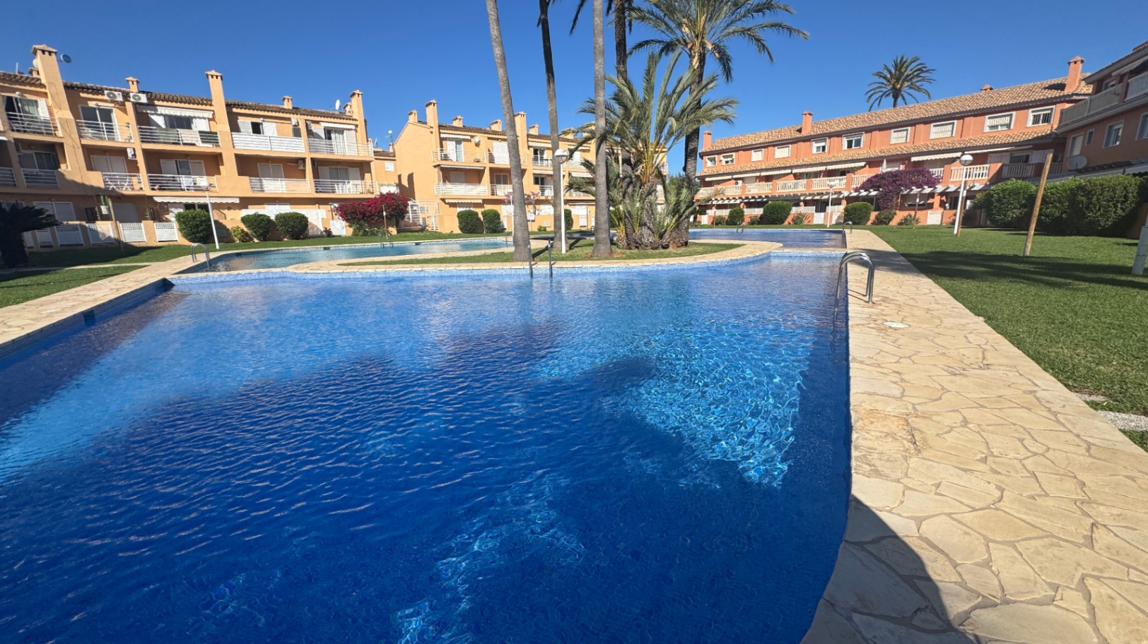 Piscina comunitaria de gresite azul con bordes de piedra natural, rodeada de zonas ajardinadas con palmeras y edificios residenciales de estilo mediterráneo.