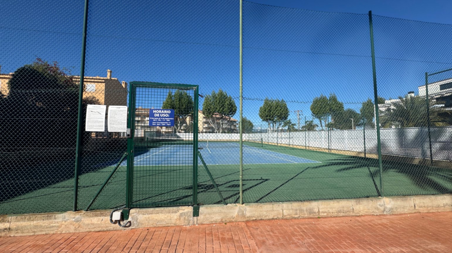 Pista de tenis exterior con superficie dura azul y verde, protegida por un vallado perimetral alto y acceso mediante puerta metálica con señalización.