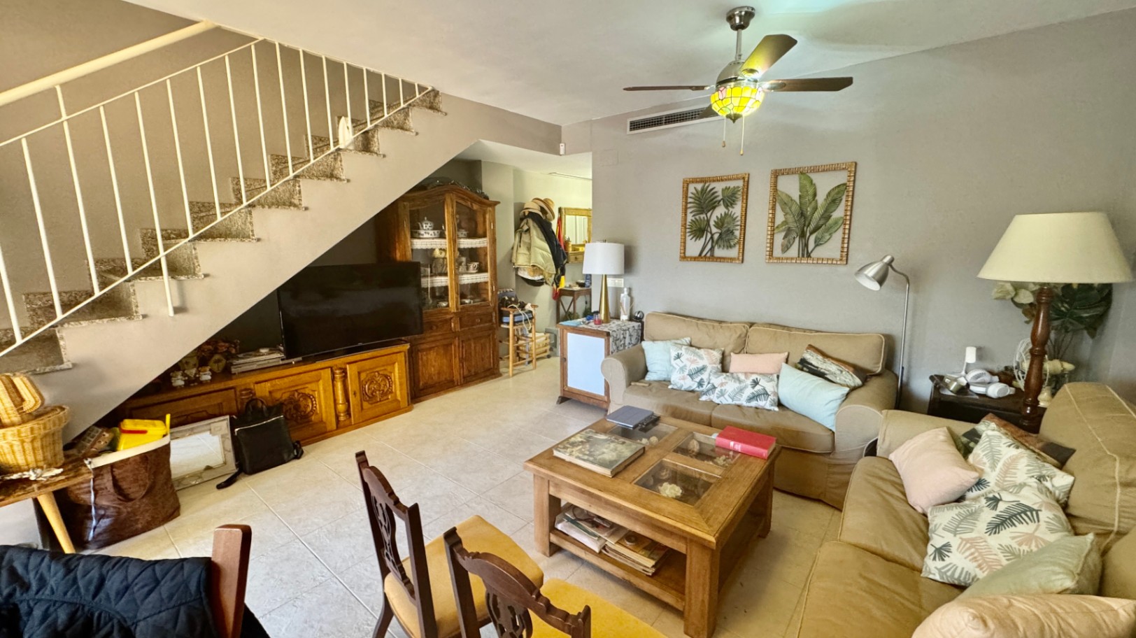 Salón con sofás beige, mesa de centro de madera y escalera con barandilla blanca. Incluye mueble de TV y vitrina de madera integrados bajo el hueco de la escalera.