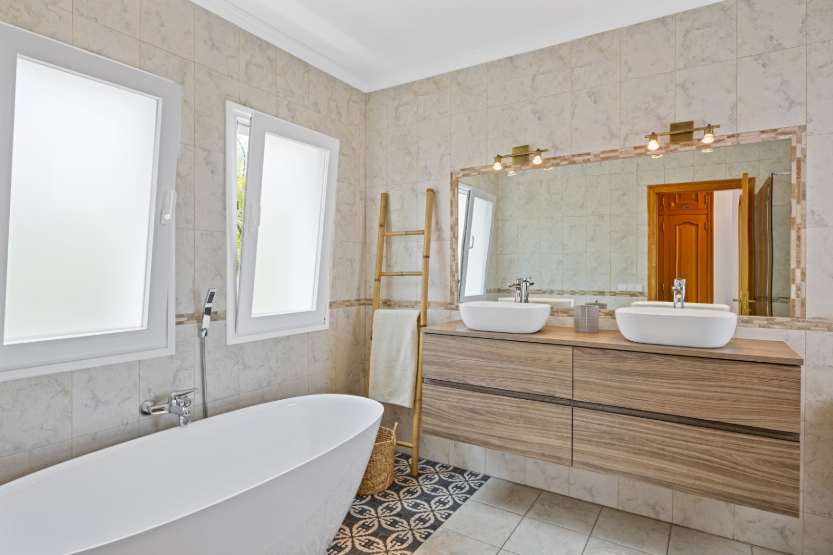Baño moderno con bañera exenta, mueble de madera con doble lavabo sobre encimera y suelo hidráulico. Espejo de gran formato con apliques de latón y ventanas de climalit.