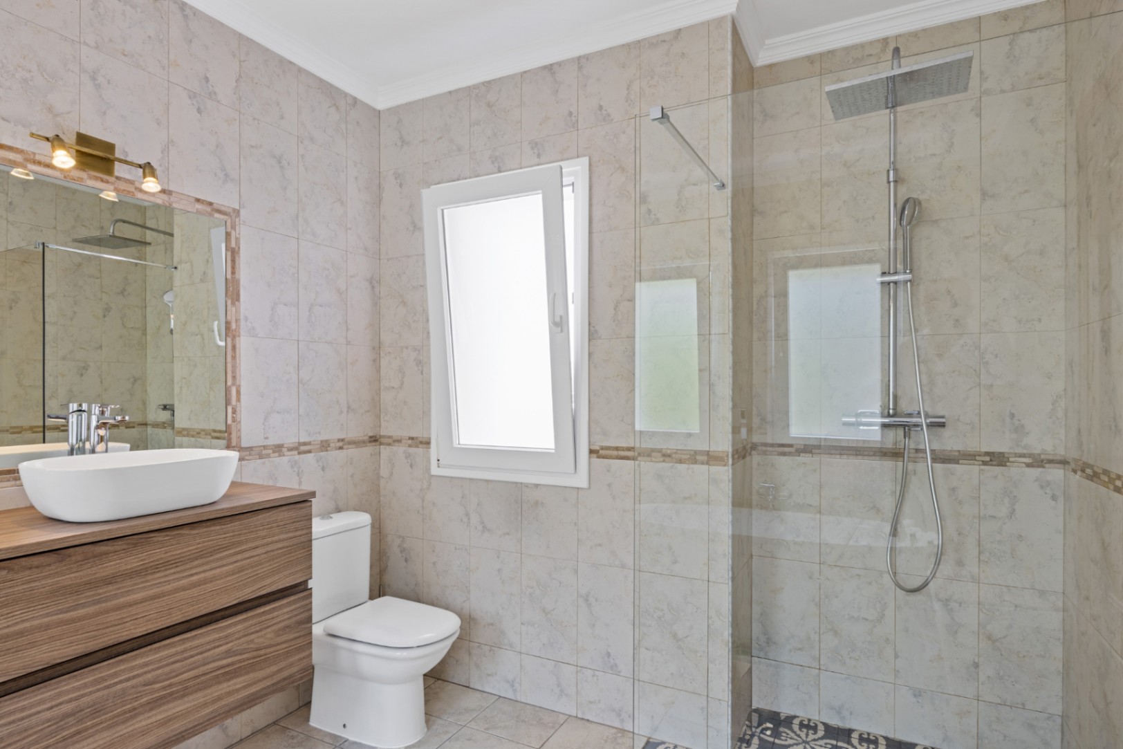 Baño moderno con ducha a ras de suelo, mampara de cristal, mueble suspendido de madera con lavabo sobre encimera y azulejos en tonos crema con luz natural.