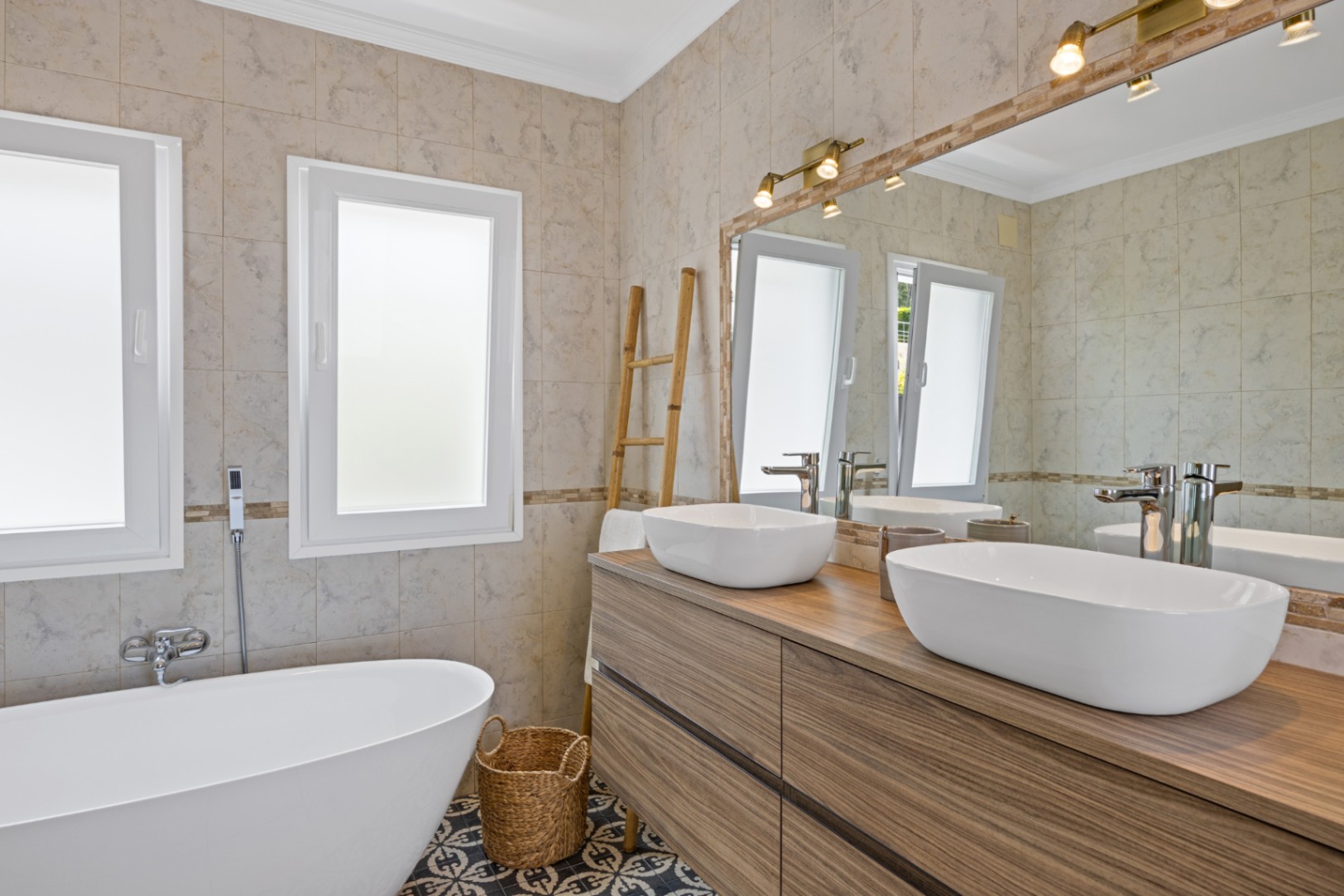Baño moderno con lavabo doble sobre mueble de madera, bañera exenta y suelo de baldosas hidráulicas decorativas, iluminado por focos y luz natural.