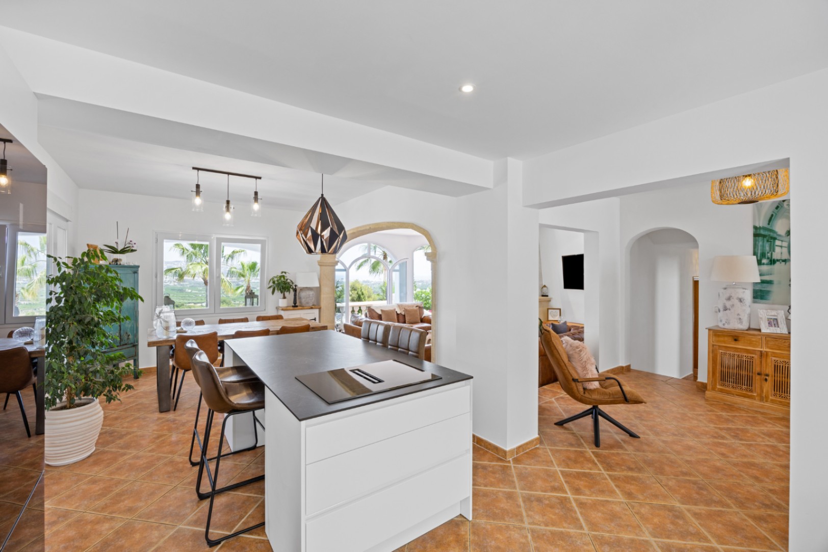 Cocina de planta abierta con isla blanca y encimera oscura, suelos de terracota y zona de comedor con vistas al exterior a través de amplios ventanales.