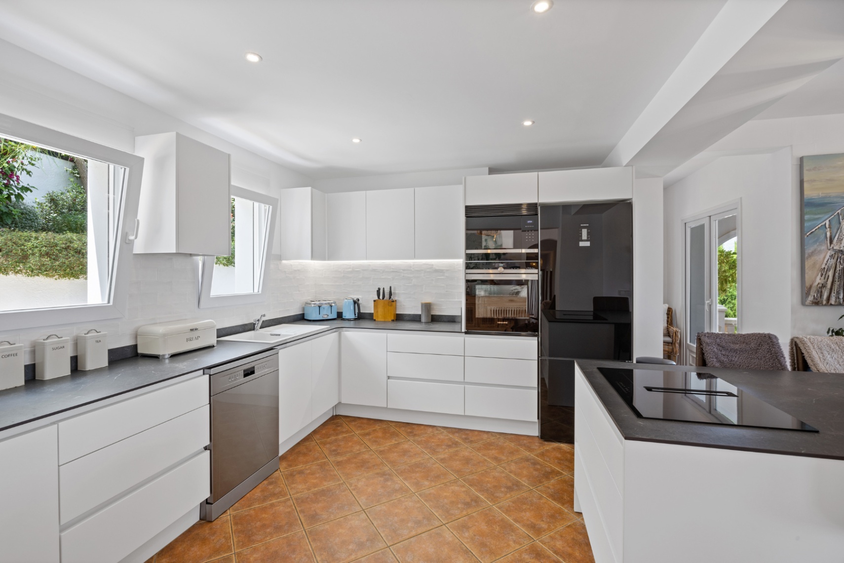 Cocina moderna con armarios blancos sin tiradores, encimeras oscuras de efecto piedra, electrodomésticos integrados y suelo cerámico de terracota.