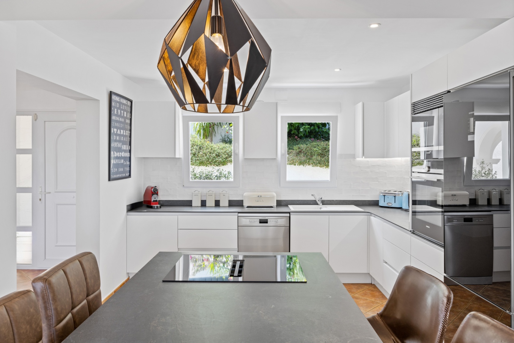 Cocina moderna con mobiliario blanco mate, encimeras de piedra oscura y una isla central con placa de inducción bajo una lámpara colgante de diseño geométrico.