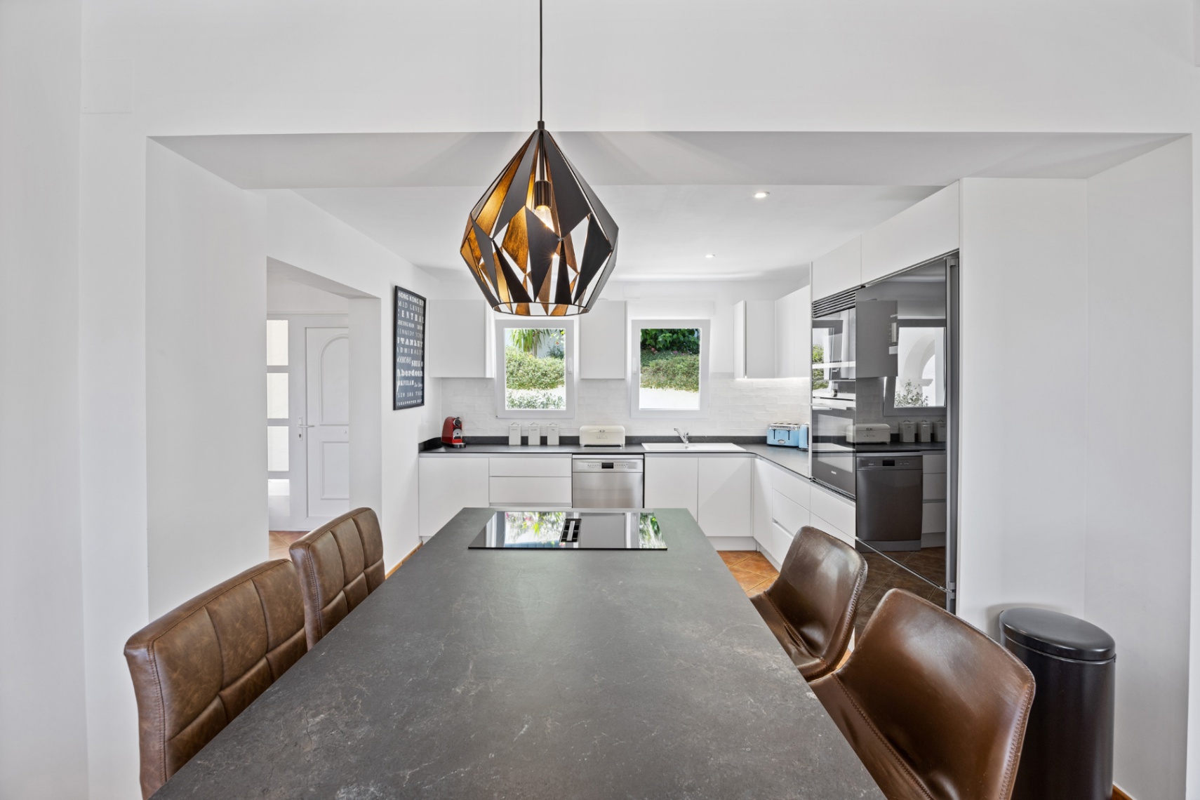 Cocina moderna con una amplia isla de piedra oscura, placa de inducción integrada, sillas de cuero marrón y armarios blancos con electrodomésticos encastrados.