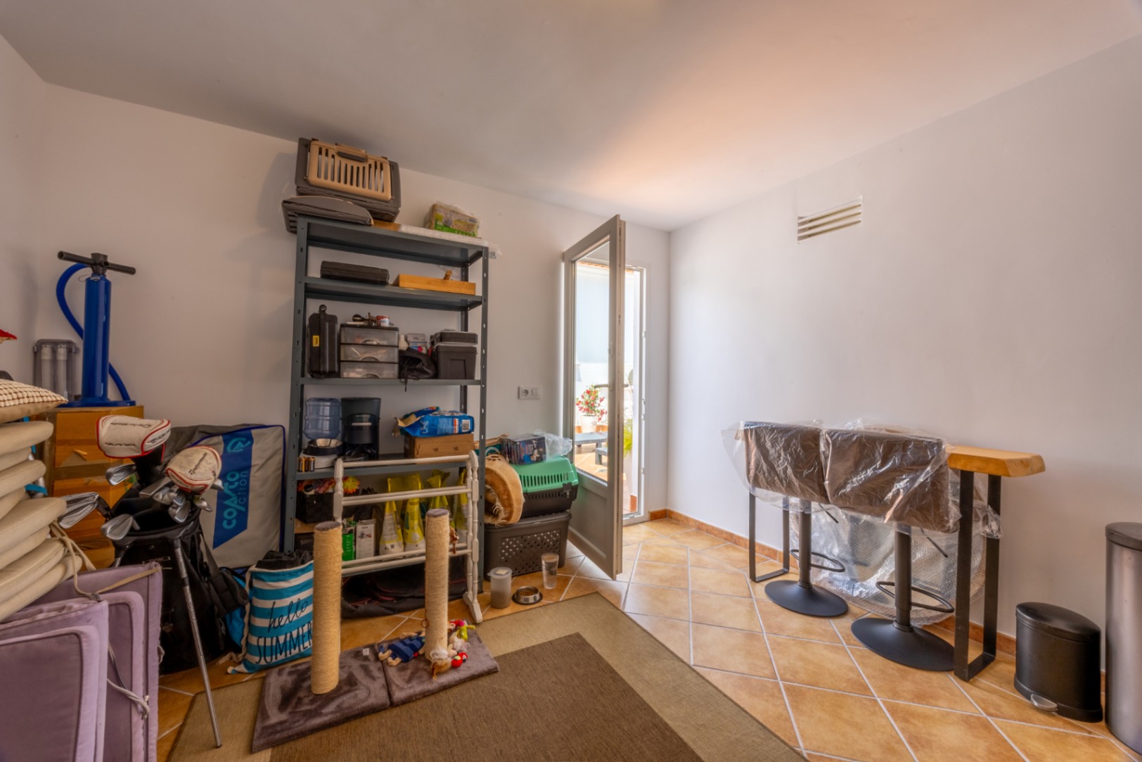 Cuarto de almacenaje con suelo de baldosas cerámicas, estantería metálica, mesa alta de madera con taburetes y acceso directo a una terraza exterior.