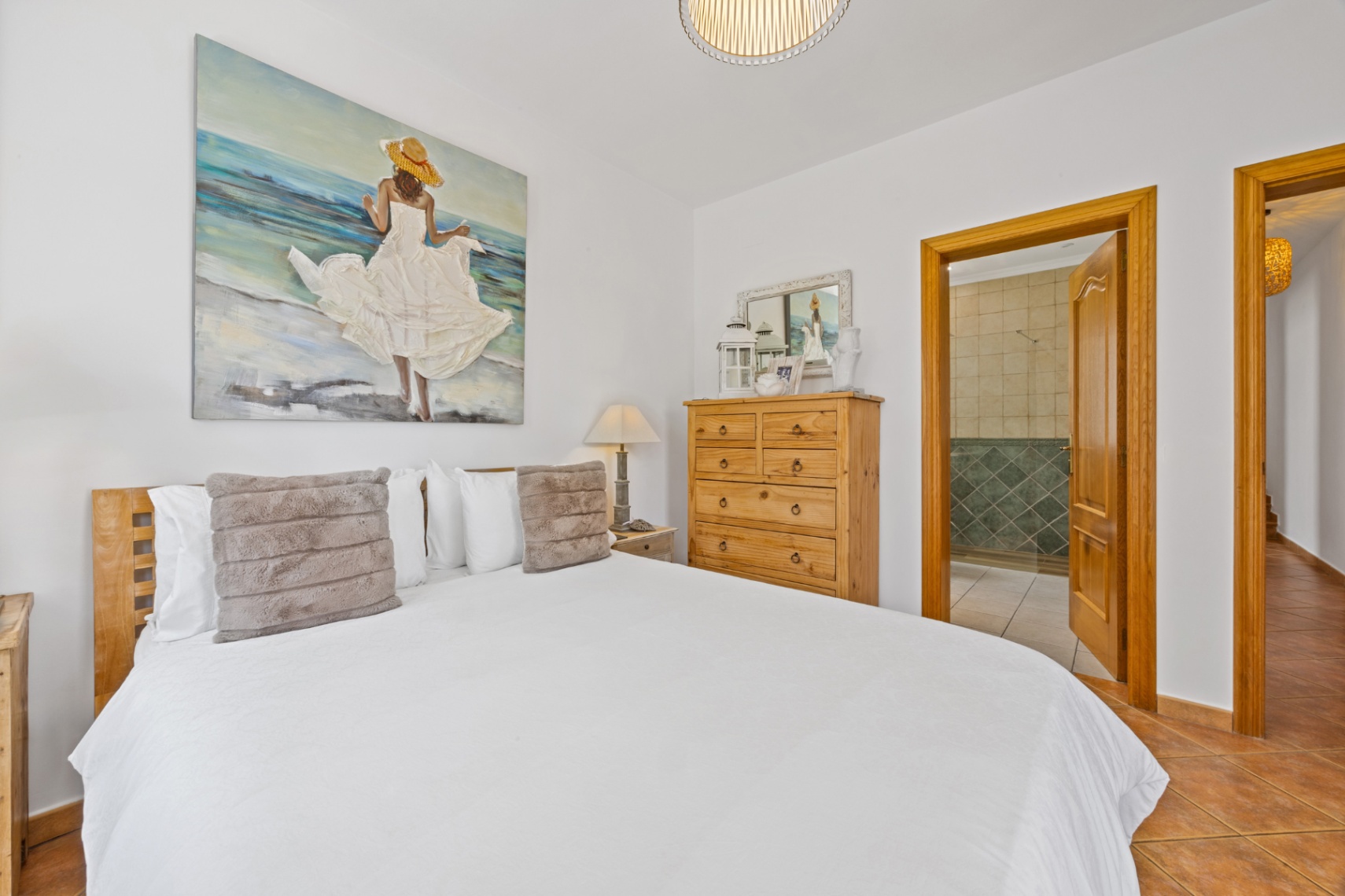 Dormitorio con cabecero de madera, gran cuadro temático y suelos de baldosa cerámica. Una puerta abierta muestra el baño en suite con azulejos verdes y ducha.