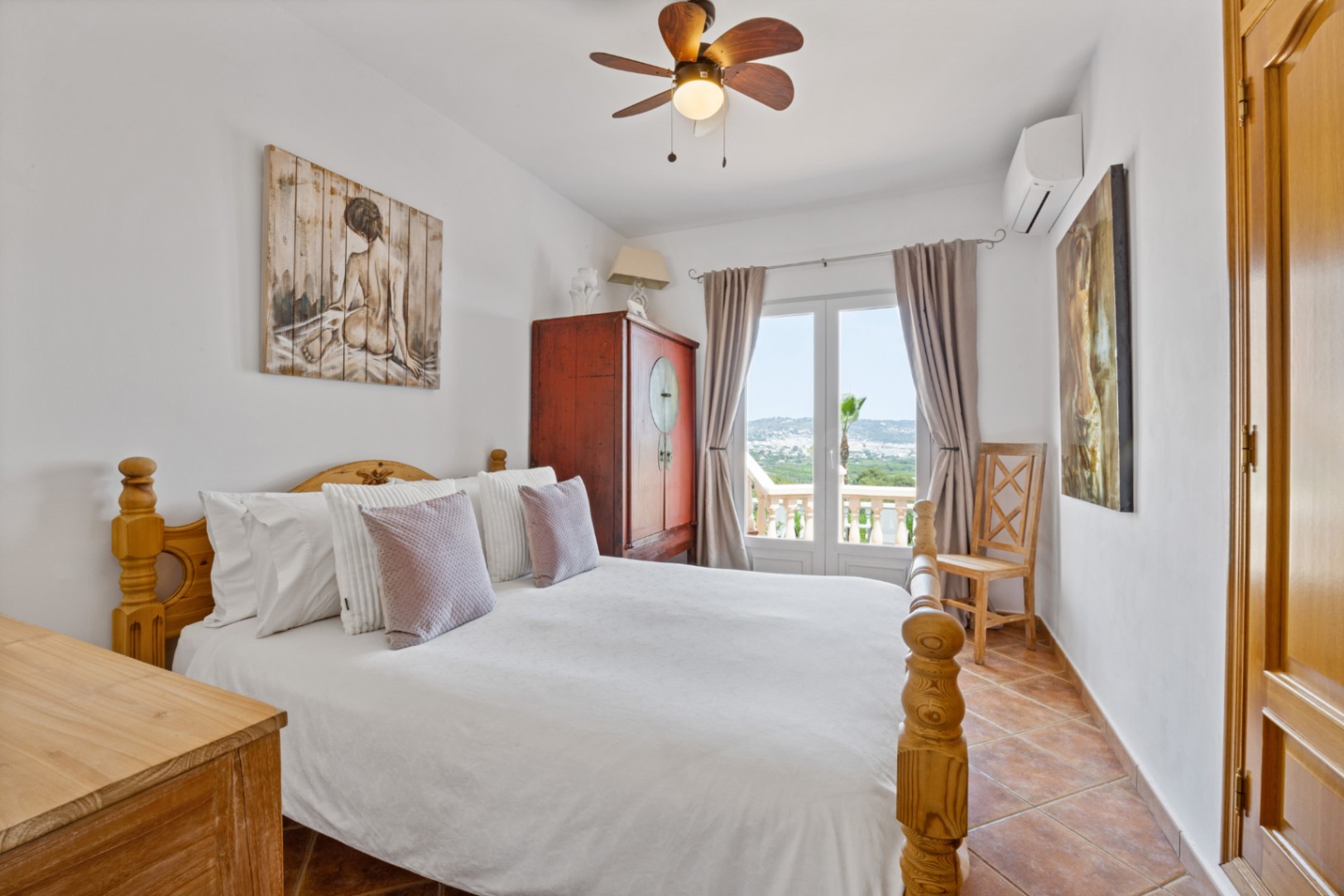 Dormitorio con cama de madera, suelo de terracota y puertas francesas que dan a un balcón con vistas. Incluye ventilador de techo y aire acondicionado.