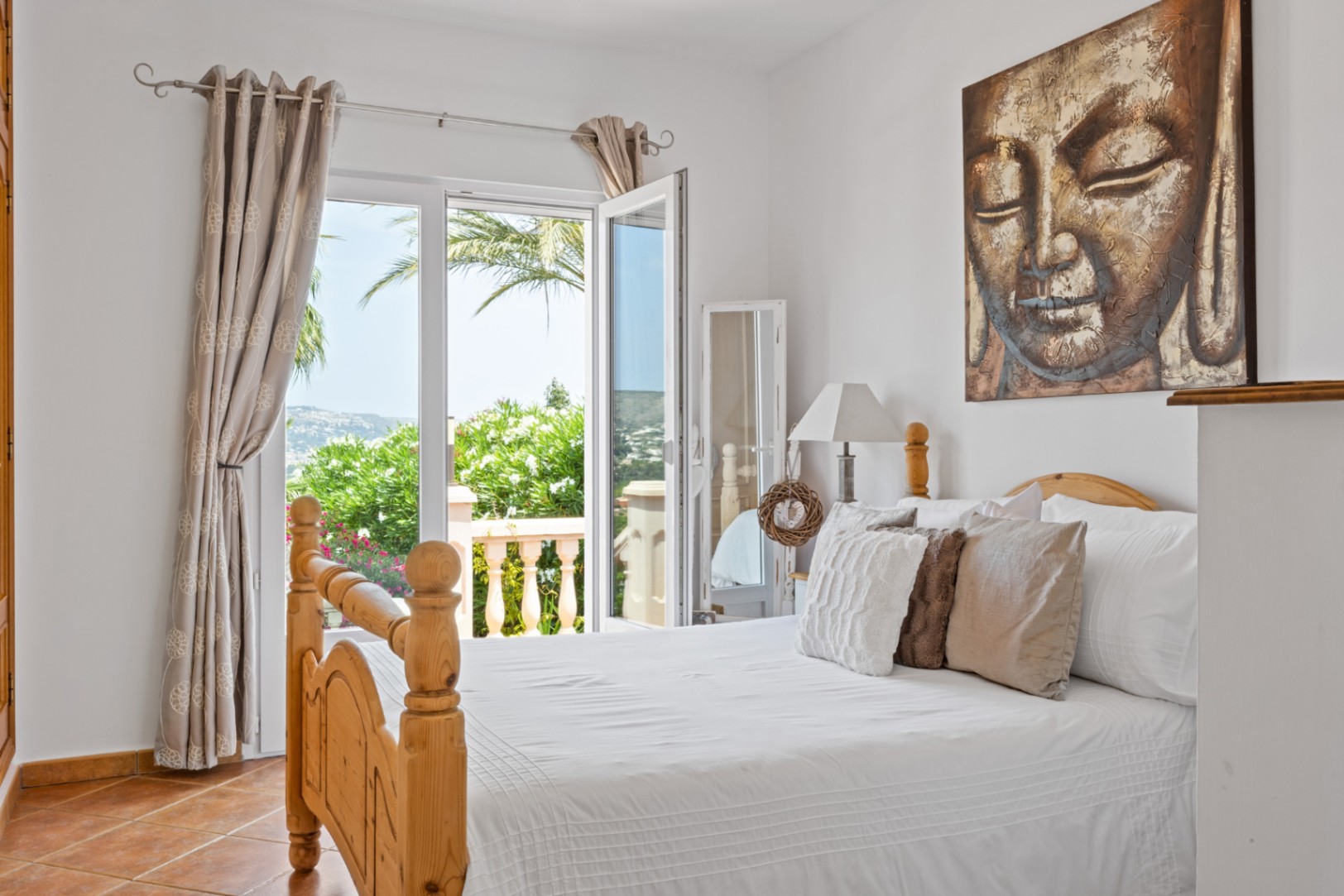Dormitorio con cama de madera, suelos de terracota y un gran cuadro de Buda. Puertas acristaladas dan acceso a un balcón con vistas al jardín mediterráneo y las colinas.